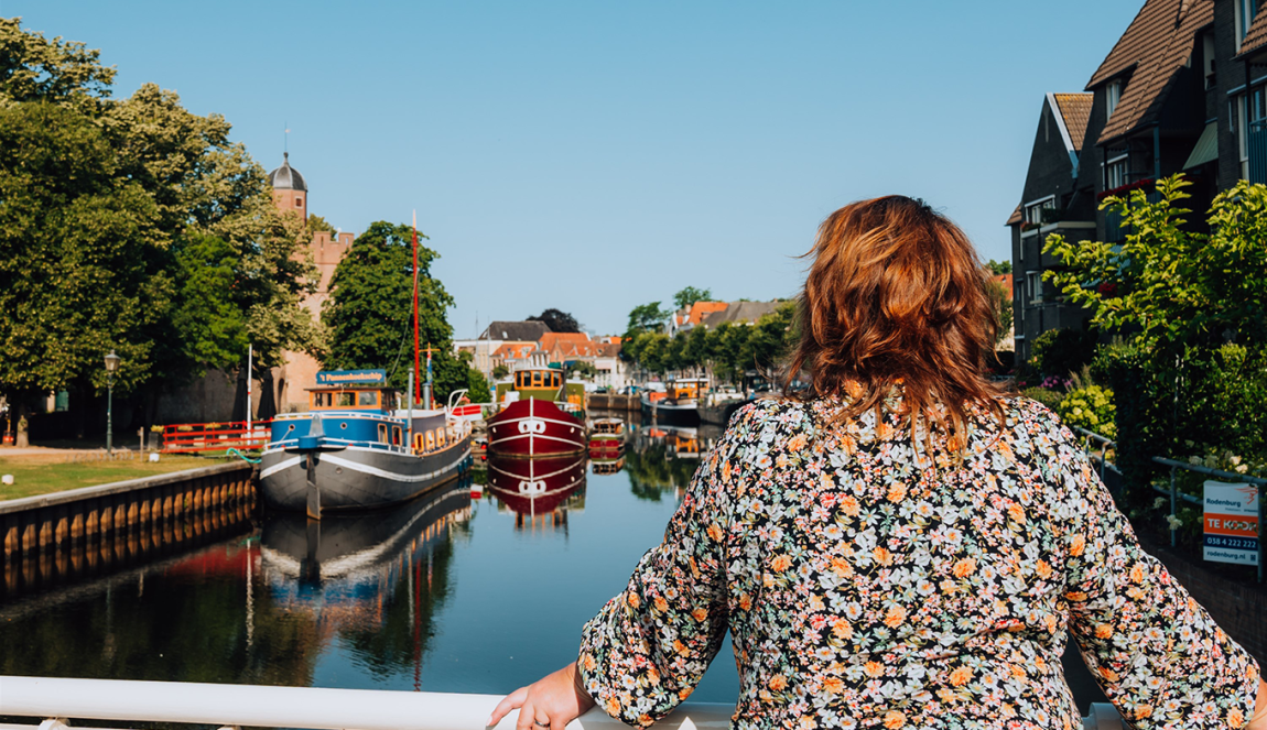 Op de rug gezien: vrouw leunt op brugreling en kijkt uit over water waarop boten aangemeerd liggen.